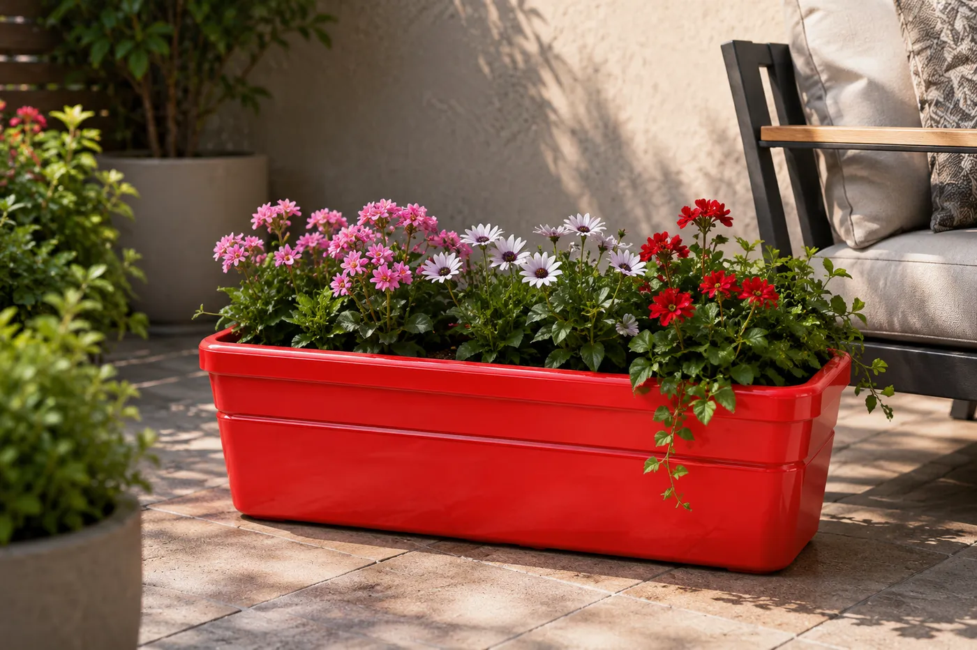 A red Fahari Rectangular trough planted with pink and white daisies, on a paved garden terrace beside a seat